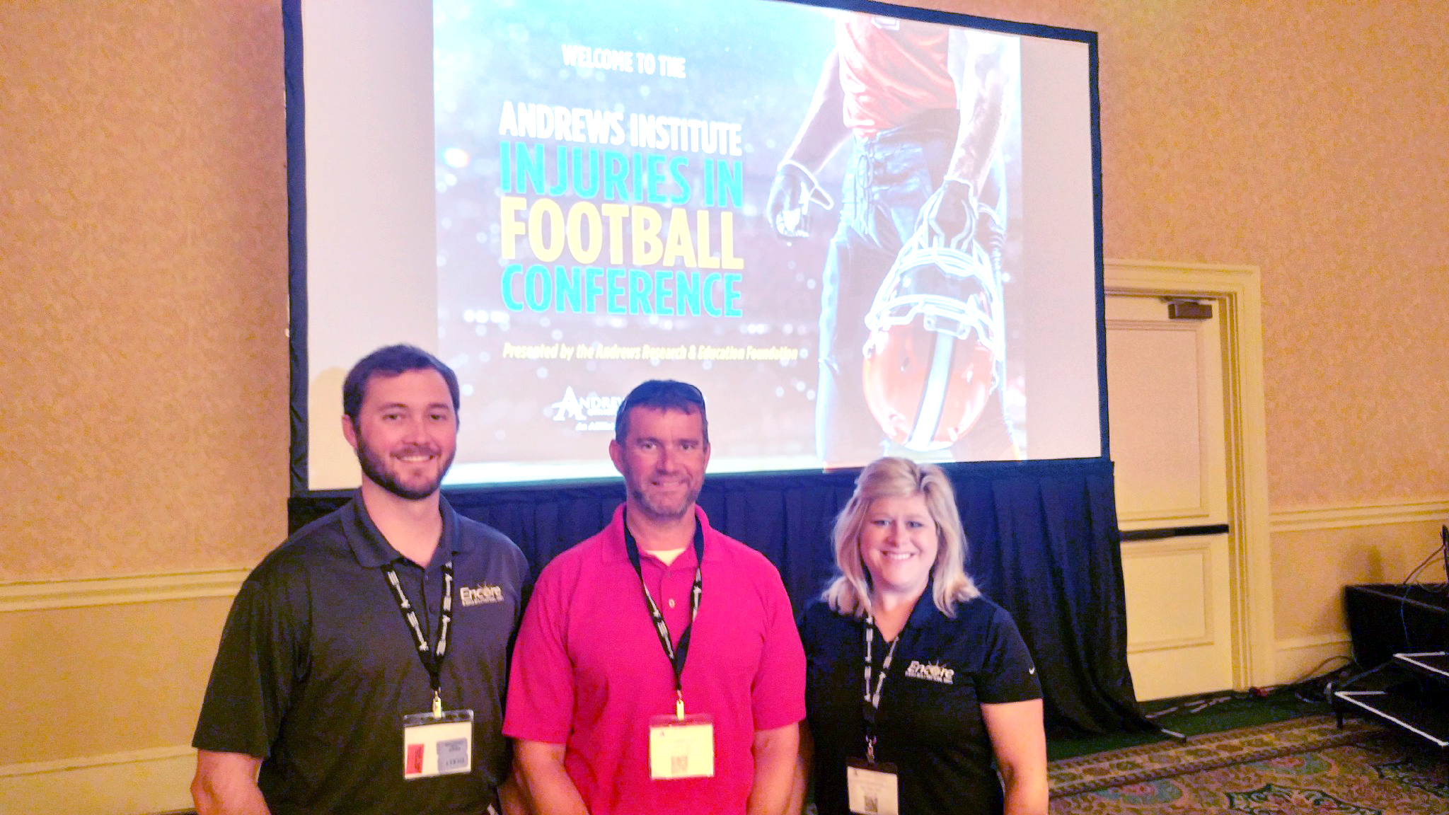 Three people from Encore Rehabilitation, Inc. pose for photo in front of movie screen displaying Andrews Institute Injuries in Football Conference