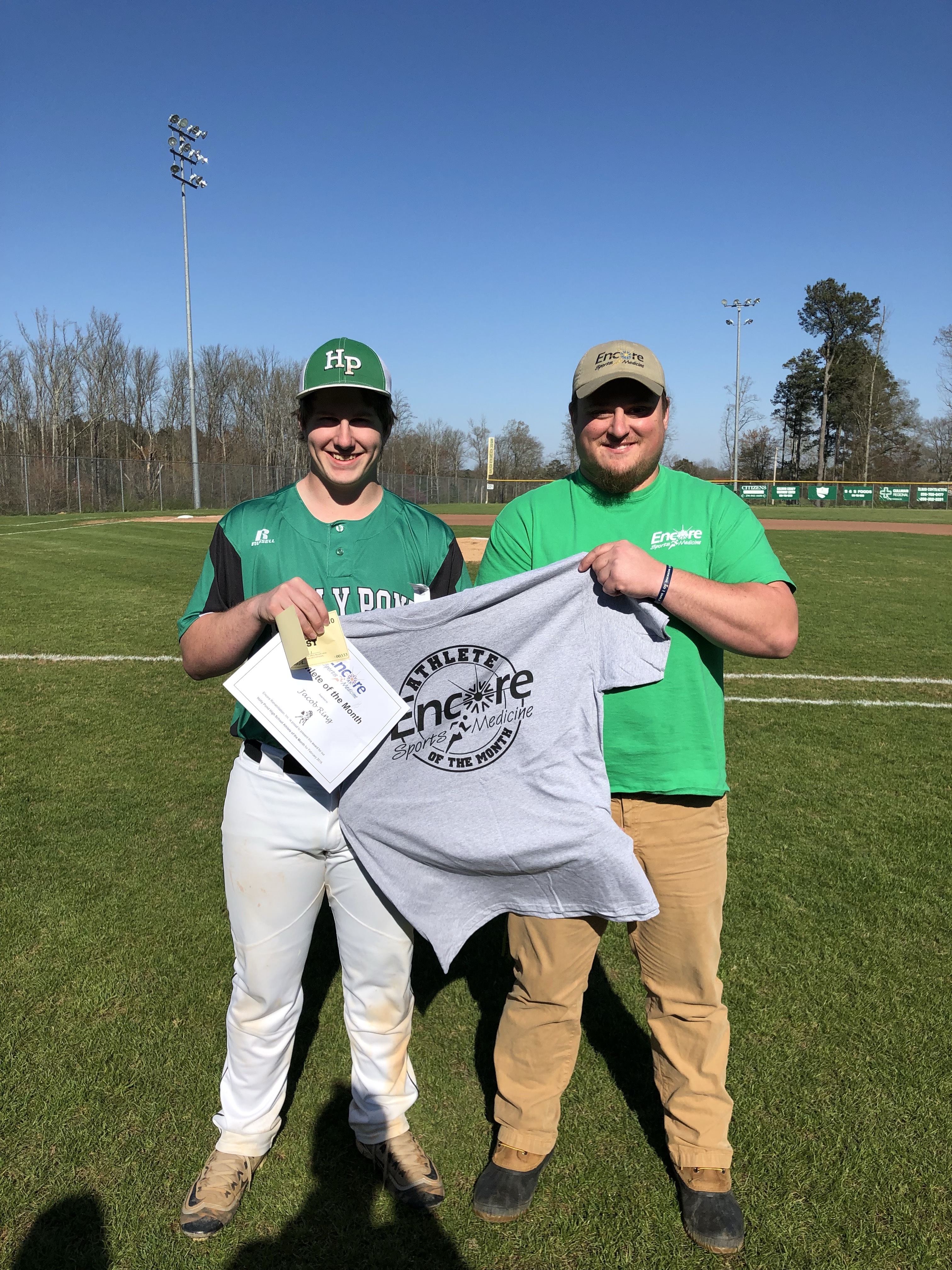 Male baseball player in uniform standing on baseball field holding a certificate and t-shirt which reads Encore Sports Medicine Athlete of the Month while male athletic trainer also helps hold shirt