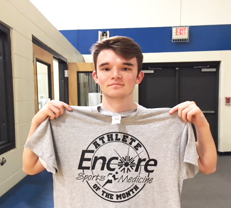 young man posing for photo and is holding up a t-shirt that read Encore Sports Medicine Athlete of the Month