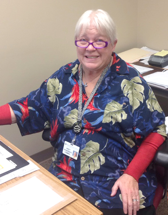 Lady in flowered shirt and glasses smiling while sitting at a desk in an office