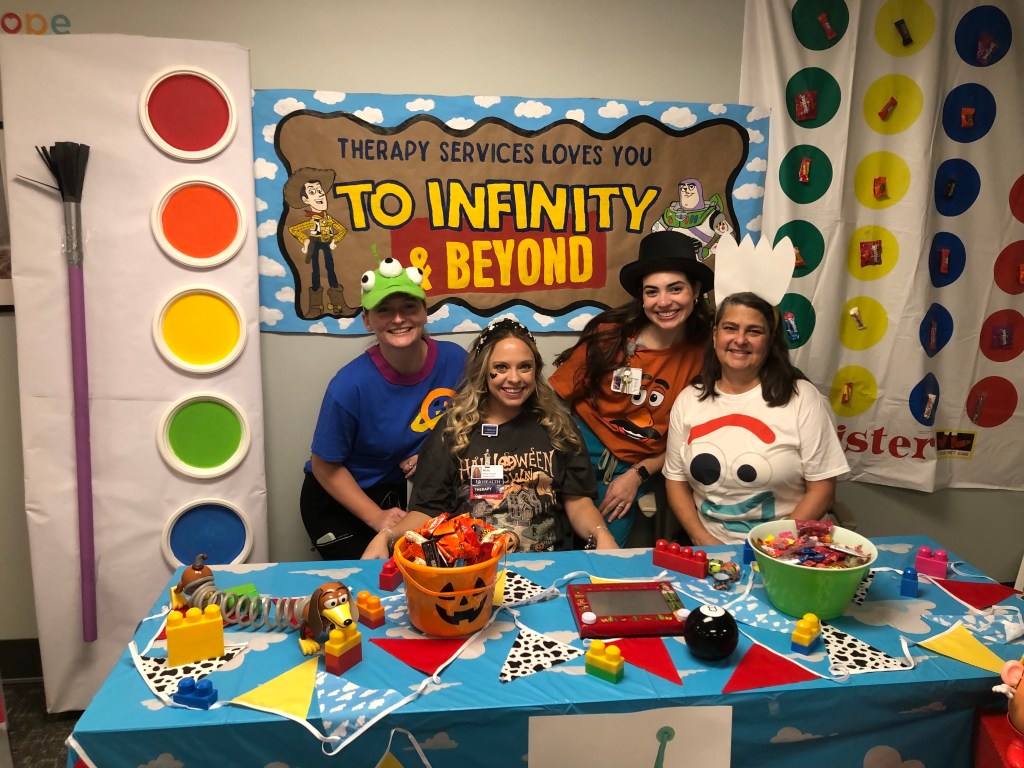 Rehab therapists dressed as characters from Toy Story stand behind a table in the hospital set up for Halloween trick-or-treating for pediatric patients. 