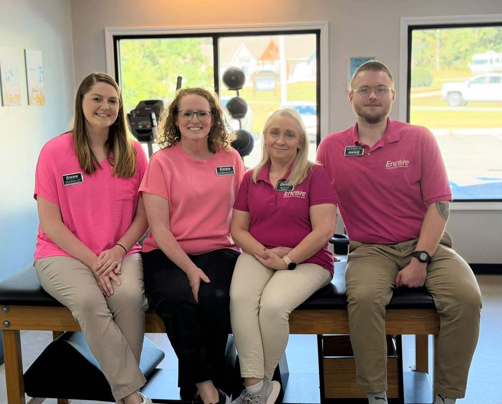 Four physical therapy staff members in matching pink shirts pose together on a treatment table in a clinic, with exercise equipment and a bright window in the background.